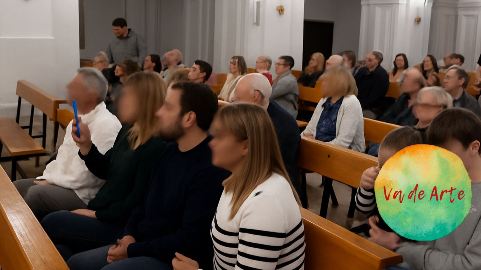 Público asistiendo a un evento cultural en un salón con bancos de madera, con el logotipo de Va de Arte superpuesto en la parte derecha.
